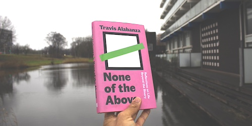 A Black hand holding a book titled 'None of the Above' in front of an Amsterdam Bijlmer lake