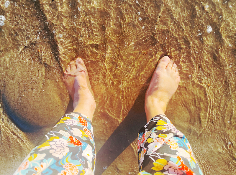 Two bare feet stand on the sand, touching the water