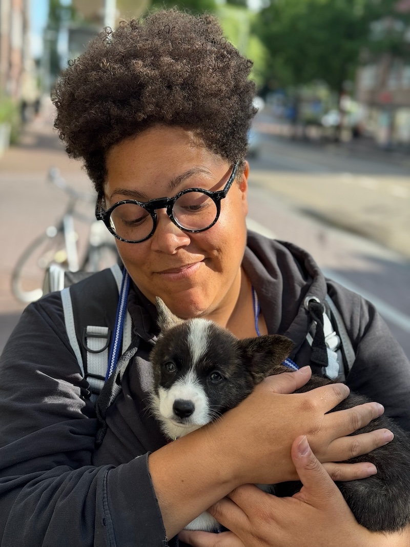 An Afropean person stands in an Amsterdam street, lovingly looking down at an eight-week-old corgi puppy in their arms