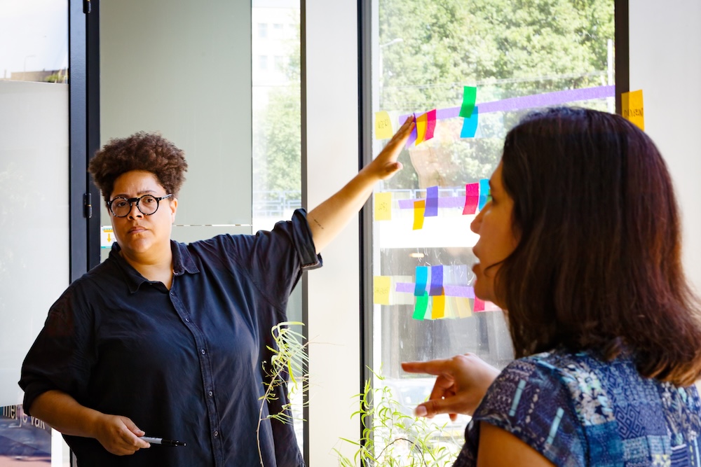 An Afropean person in a black shirt points at a post-it on a window, looking at their coworker explain a concept