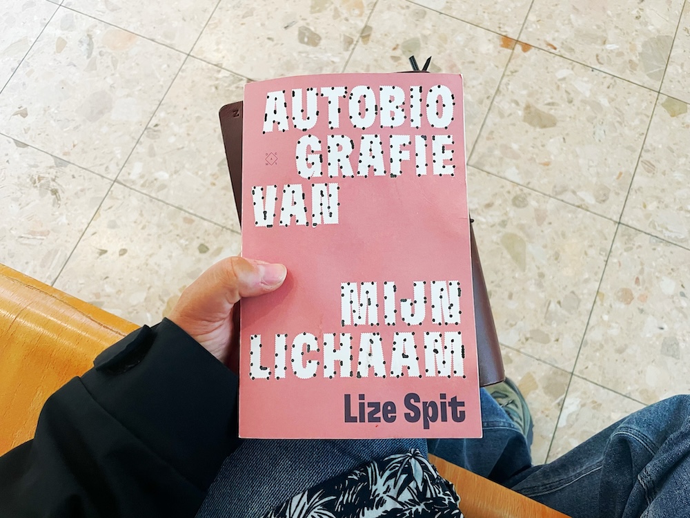 A person holds a book in their lap titled 'Autobiography of my body', by Flemish author Lize Spit