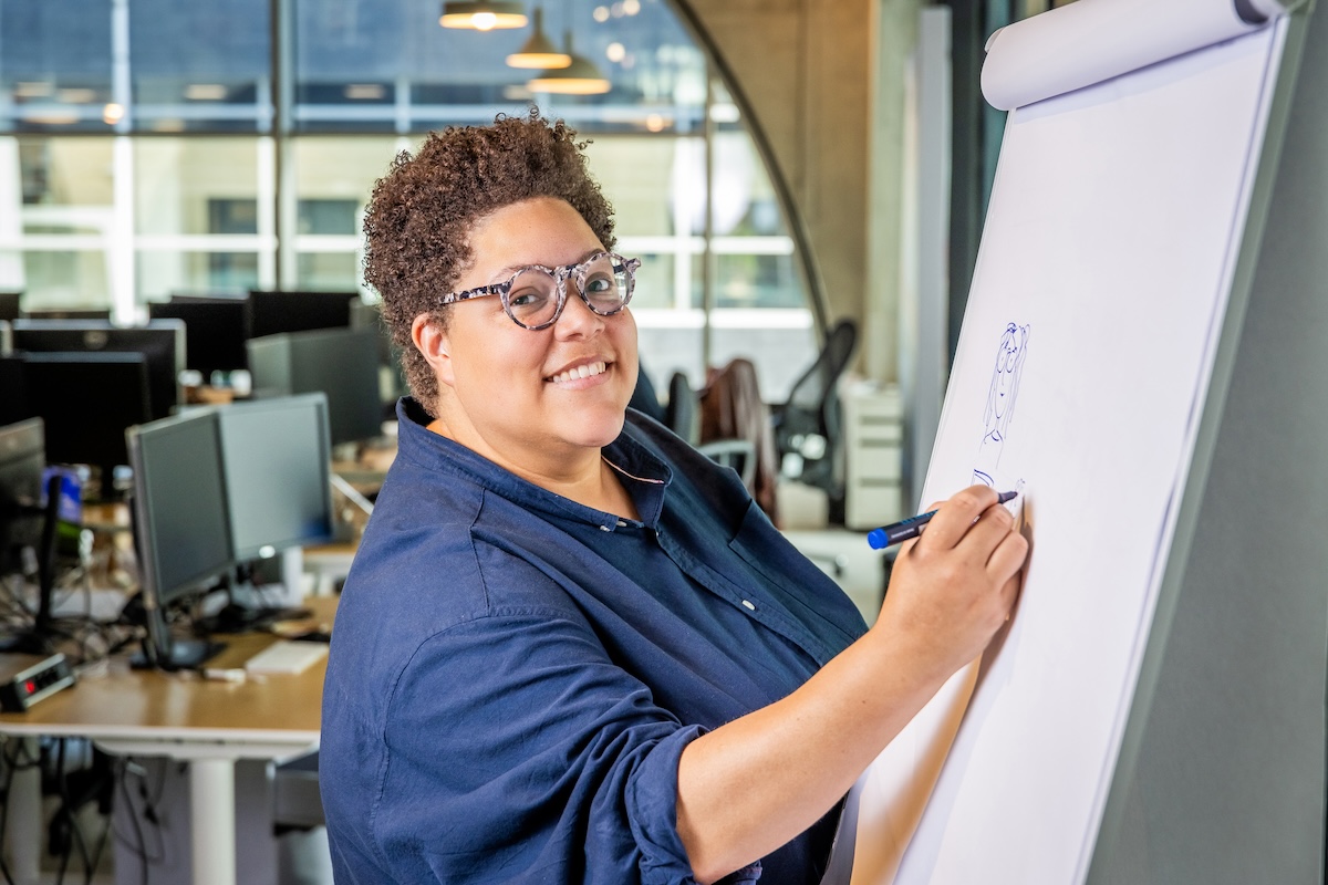 A Black person at the office smiling at the camera while doodling on a flipchart